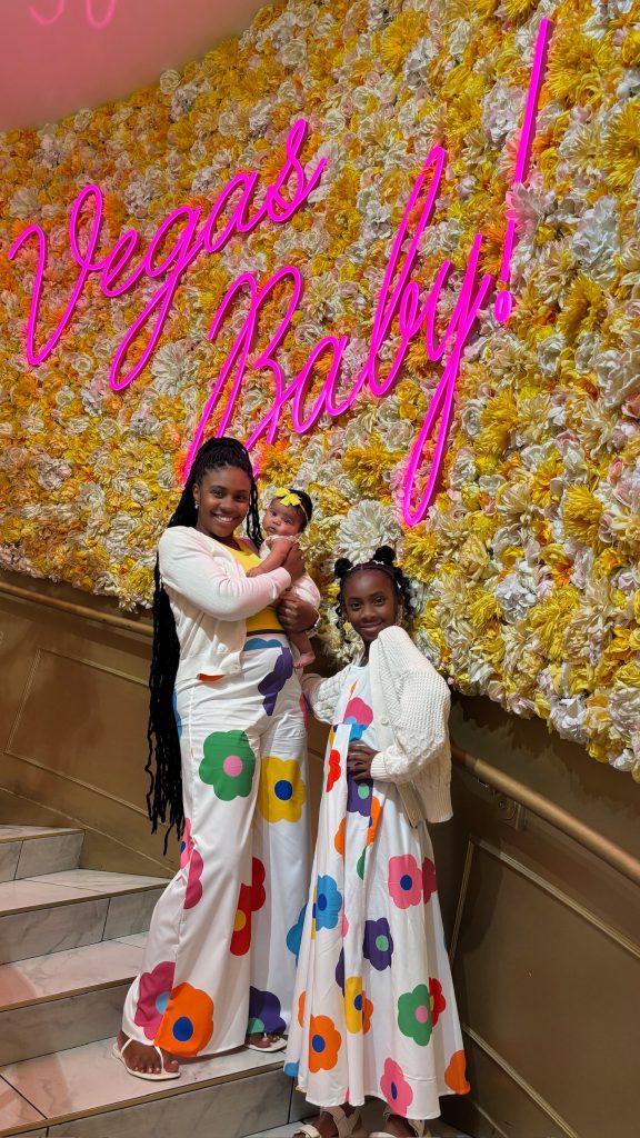 Sucoyia posing with her daughters and baby in coordinated floral outfits in front of a neon “Vegas Baby!” flower wall.