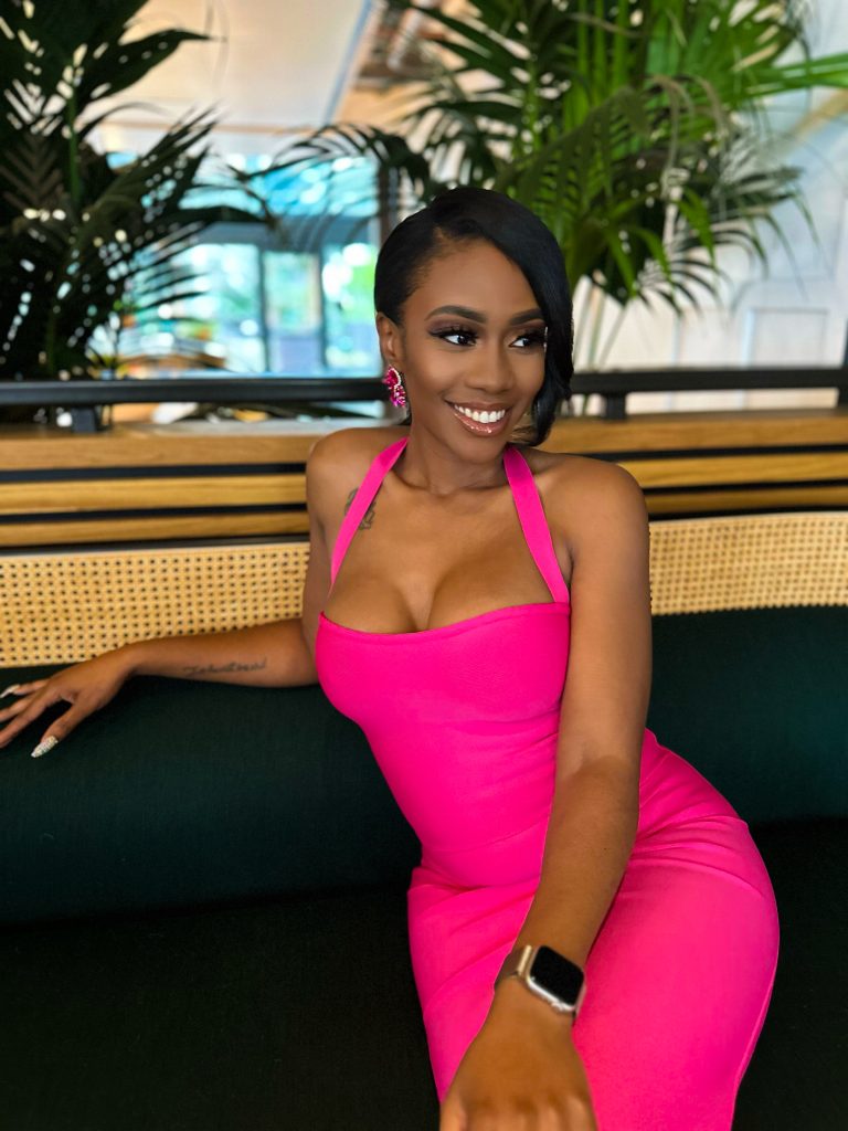 Sucoyia smiling in a bright pink dress while seated in a modern upscale restaurant setting.